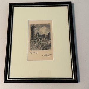 May include: A framed black and white etching of a castle scene with a tall tower, featuring the text "ROTHENBURG" at the top and a signature at the bottom. The artwork is set within a cream-colored mat and a dark brown frame with silver trim.