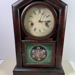 May include: Antique wooden clock with a dark brown finish. The clock face has Roman numerals and hands. Below the clock face is a decorative panel with a green background and a circular design. A small key rests in front of the clock.