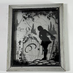 May include: A framed silhouette art piece with a silver-colored frame. The artwork depicts a scene with a figure leaning on a cane, a gate, and a house. The art is black and white.