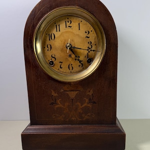 May include: Antique wooden Seth Thomas clock with a gold-tone clock face. The clock has a dark brown wooden case with an arched top and a decorative floral inlay. The clock face features black numerals and hands.