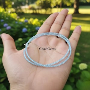Op de afbeelding: Een delicate, lichtblauwe kralenketting wordt op een open hand getoond. De ketting is gemaakt van kleine, ronde kralen en is om de hand gewikkeld. De tekst "ChaviGems" is zichtbaar.