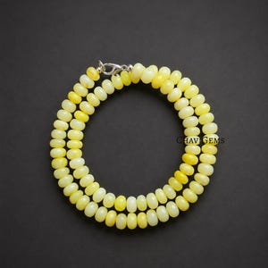 May include: A necklace composed of light yellow, oval-shaped beads. The beads are strung together, forming a double strand. A silver-colored clasp secures the necklace. The background is a solid black.