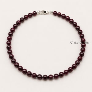 May include: A necklace composed of small, round, deep red beads. The beads are strung together, forming a complete circle. A silver-colored clasp secures the necklace. The text "ChaviGems" is visible on the right side of the image.