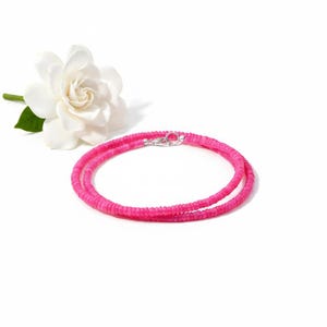 Puede incluir: Un collar de cuentas rosa brillante con cierre plateado, sobre fondo blanco. Una flor de gardenia blanca con hojas verdes en el fondo. Un accesorio vibrante.