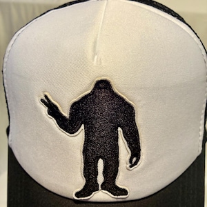 Pode incluir: Boné de caminhoneiro preto e branco com uma silhueta preta de Bigfoot fazendo o sinal de paz. O painel frontal é branco, com um patch preto de Bigfoot. A aba e a parte de trás em malha são pretas, e o boné tem um botão preto na parte superior.