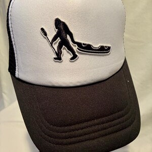 Op de afbeelding: Zwarte en witte truckerpet met een Bigfoot-silhouet dat een peddel en een kajak draagt. De pet heeft een wit voorpaneel met het geborduurde ontwerp, een zwarte klep en een zwarte mesh-achterkant.