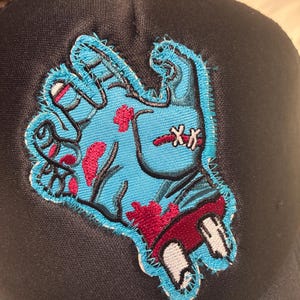 以下が含まれることがあります： ゾンビの手のパッチが付いた黒いトラッカーハット。パッチは、赤い血のディテールとステッチが施された水色の手を示しています。帽子は湾曲したつばとメッシュの背面を備えています。