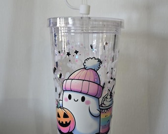 May include: Clear plastic tumbler with a Halloween ghost design. The ghost wears a pink beanie and holds a pumpkin and a rainbow drink. The lid has a ghost-shaped straw topper. The tumbler is filled with glitter and star-shaped confetti.