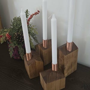 May include: Four white taper candles with copper-coloured bases sit on wooden block candle holders. The candle holders are arranged on a dark wooden surface. A small decorative plant is in the background.