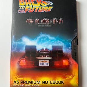 May include: A5 premium notebook with 240 lined pages. The cover displays the "Back to the Future" logo and a DeLorean car illustration. The destination time reads November 5, 1955, at 01:21 AM. The notebook is black with orange and blue accents.