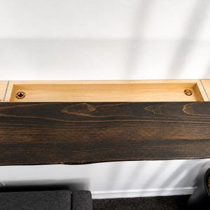 May include: A wall-mounted floating shelf featuring a dark brown stained wood top with a natural live edge and a light-coloured wooden storage compartment underneath. The interior of the storage tray includes two circular branded logos on the left and right.