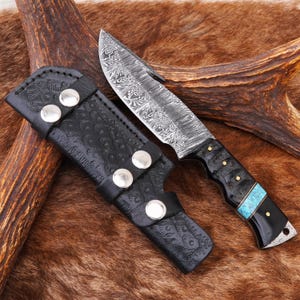 May include: A Damascus steel hunting knife with a black handle, turquoise inlay, and a black leather sheath. The blade has a unique pattern. The sheath is embossed with a pattern and secured with silver-colored rivets.