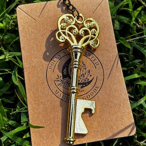 May include: A gold-colored metal key-shaped bottle opener with an ornate, filigree-style bow and a functional bit. The opener is attached to a dark metal jump ring and mounted on a rectangular brown card featuring the text Dolly's Enchanted Goods.