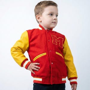May include: A red and yellow varsity jacket with the letter "M" on the chest. The jacket has yellow sleeves and white and yellow striped cuffs and hem. The jacket is worn by a young child.