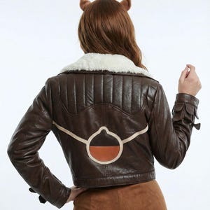 May include: A brown leather jacket with a cream shearling collar. The back has a distinctive tan and brown patch. The jacket is cropped and worn with brown trousers. The jacket has a classic aviator style, with a vintage aesthetic.