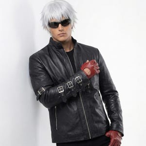 Op de afbeelding: Een zwarte leren jas met ritssluiting, gedragen met een zwarte broek en rode vingerloze handschoenen. De jas heeft zilveren gespen op de mouwen. De persoon draagt een zonnebril en heeft wit haar.