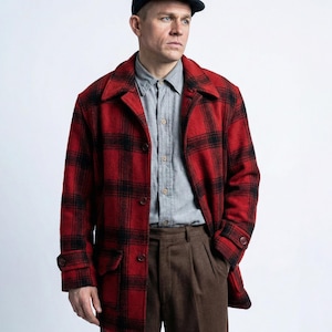 May include: A red and black checked wool coat with a button closure, worn over a grey button-down shirt and brown trousers. A dark blue cap completes the outfit. The coat has a classic, outdoorsy style.