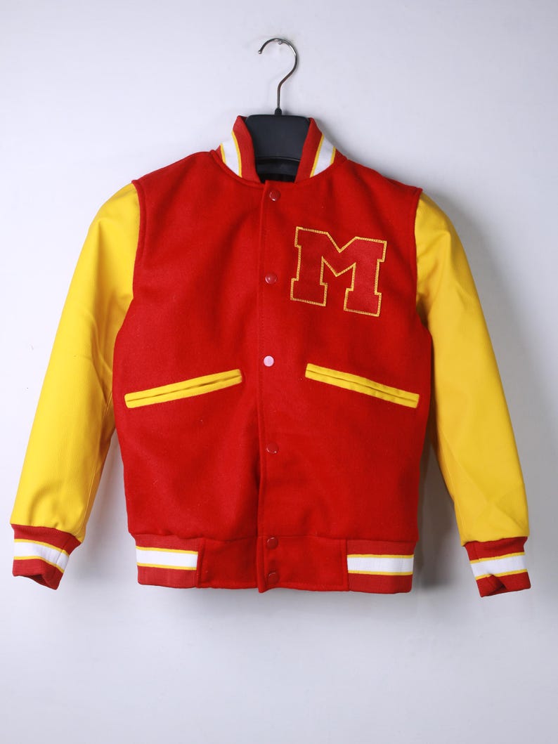 May include: A red and yellow varsity jacket with the letter "M" on the chest. The jacket has yellow sleeves, white and yellow striped cuffs and a white collar. The jacket is hanging on a black hanger.