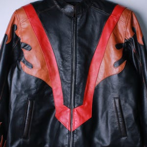 K&ouml;nnte beinhalten: Schwarze Lederjacke mit Stehkragen und durchgehendem Rei&szlig;verschluss. Die Jacke hat rote und braune flammenartige Designs auf den Schultern und der Brust. Die Jacke hat zwei Vordertaschen.