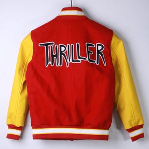 May include: Red and yellow varsity jacket with the word "THRILLER" embroidered on the back in black and white. The jacket has yellow sleeves, a red body, and white and red striped cuffs and collar.