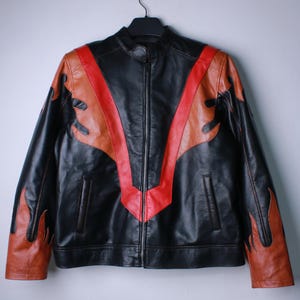 K&ouml;nnte beinhalten: Schwarze Lederjacke mit durchgehendem Rei&szlig;verschluss, mit einem roten und braunen Flammendesign auf der Vorderseite und den &Auml;rmeln. Die Jacke h&auml;ngt an einem schwarzen Kleiderb&uuml;gel vor einem schlichten Hintergrund.