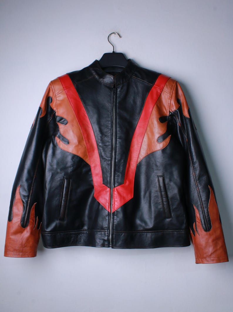 K&ouml;nnte beinhalten: Schwarze Lederjacke mit durchgehendem Rei&szlig;verschluss, mit einem roten und braunen Flammendesign auf der Vorderseite und den &Auml;rmeln. Die Jacke h&auml;ngt an einem schwarzen Kleiderb&uuml;gel vor einem schlichten Hintergrund.