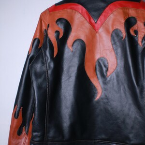 K&ouml;nnte beinhalten: Schwarze Lederjacke mit orangefarbenen und roten Flammenakzenten. Die Jacke hat einen klassischen Kragen und eine taillierte Passform. Die Flammen sind auf Schultern und R&uuml;cken aufgen&auml;ht und erzeugen einen auff&auml;lligen Look.