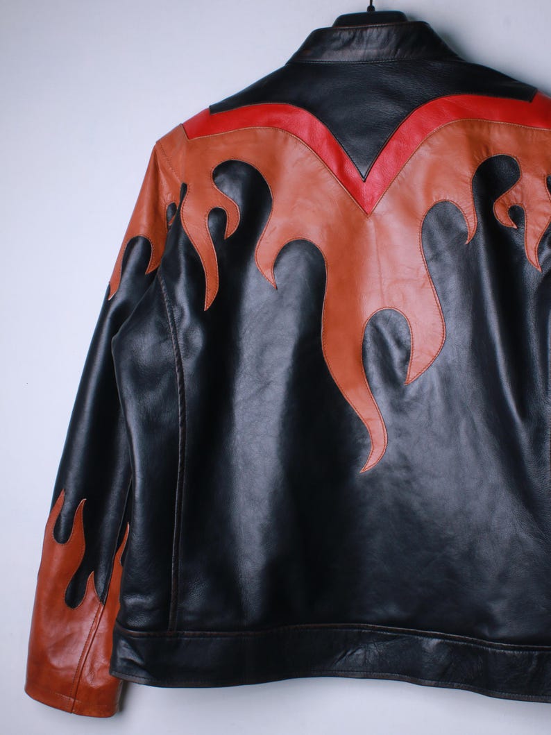 K&ouml;nnte beinhalten: Schwarze Lederjacke mit orangefarbenen und roten Flammenakzenten. Die Jacke hat einen klassischen Kragen und eine taillierte Passform. Die Flammen sind auf Schultern und R&uuml;cken aufgen&auml;ht und erzeugen einen auff&auml;lligen Look.