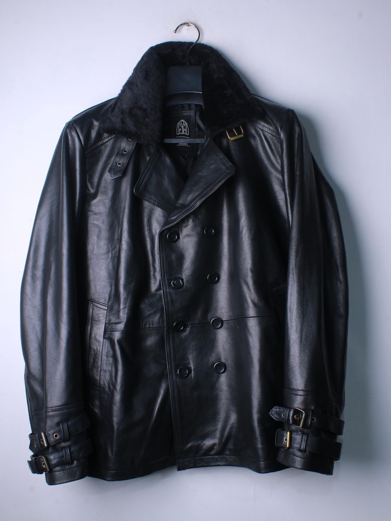 May include: Black leather jacket with a faux fur collar, double-breasted front, and buckle details on the sleeves. The jacket is hanging on a hanger against a plain background. The jacket has a classic, timeless design.