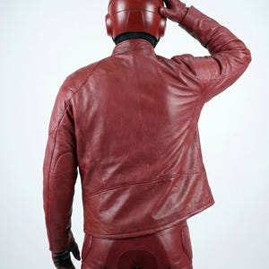 以下が含まれることがあります： 赤い革のジャケット、パンツ、手袋、ヘルメットを着用した人物。 衣装は濃い赤色です。人物はカメラから背を向けており、頭に手を置いています。