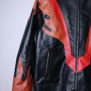 K&ouml;nnte beinhalten: Schwarze Lederjacke mit Flammen- und Chevron-Designs in Rot und Braun. Die Jacke hat einen Rei&szlig;verschluss vorne und eine kleine Tasche. Die Flammen befinden sich auf den &Auml;rmeln und das Chevron-Design auf der Brust.