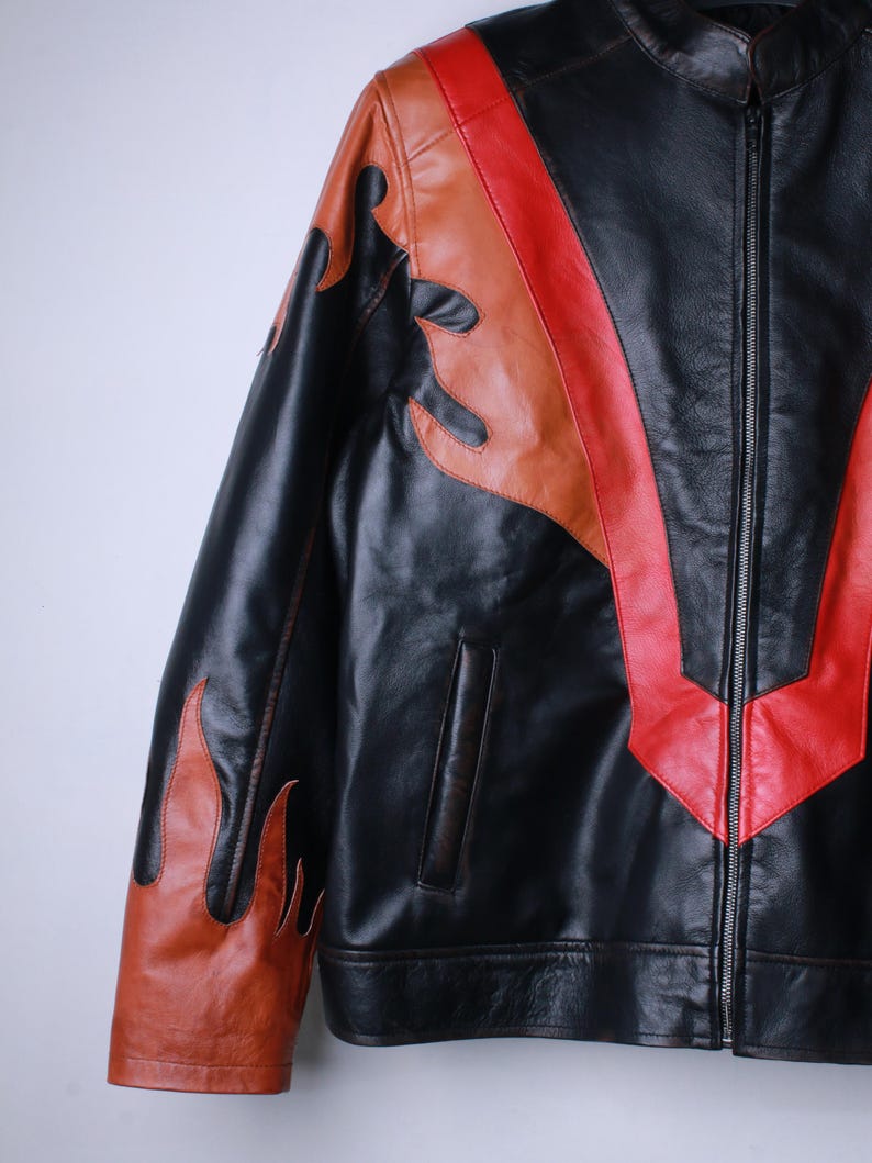 K&ouml;nnte beinhalten: Schwarze Lederjacke mit Flammen- und Chevron-Designs in Rot und Braun. Die Jacke hat einen Rei&szlig;verschluss vorne und eine kleine Tasche. Die Flammen befinden sich auf den &Auml;rmeln und das Chevron-Design auf der Brust.