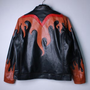 K&ouml;nnte beinhalten: Schwarze Lederjacke mit orangefarbenen und roten Flammenakzenten. Die Jacke hat einen klassischen Biker-Stil mit Stehkragen und durchgehendem Rei&szlig;verschluss. Das Flammendesign befindet sich auf Schultern, &Auml;rmeln und R&uuml;cken.