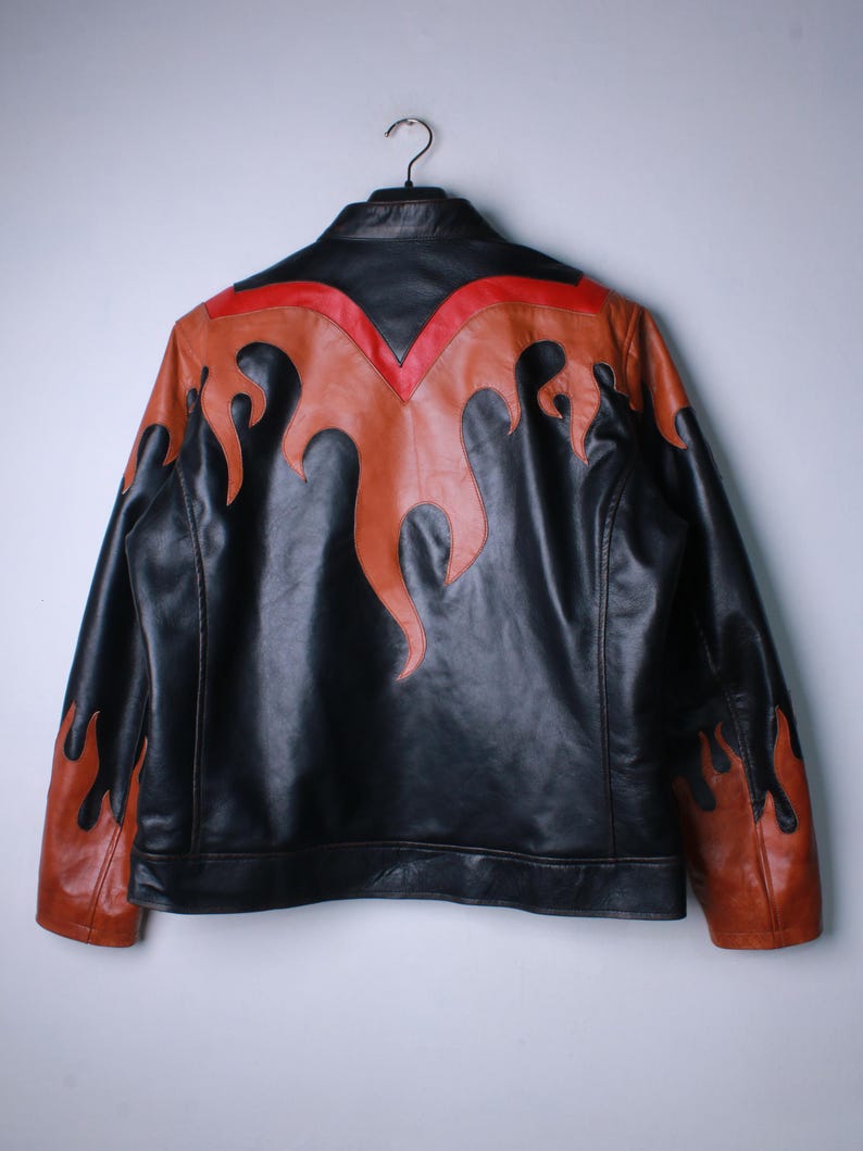 K&ouml;nnte beinhalten: Schwarze Lederjacke mit orangefarbenen und roten Flammenakzenten. Die Jacke hat einen klassischen Biker-Stil mit Stehkragen und durchgehendem Rei&szlig;verschluss. Das Flammendesign befindet sich auf Schultern, &Auml;rmeln und R&uuml;cken.