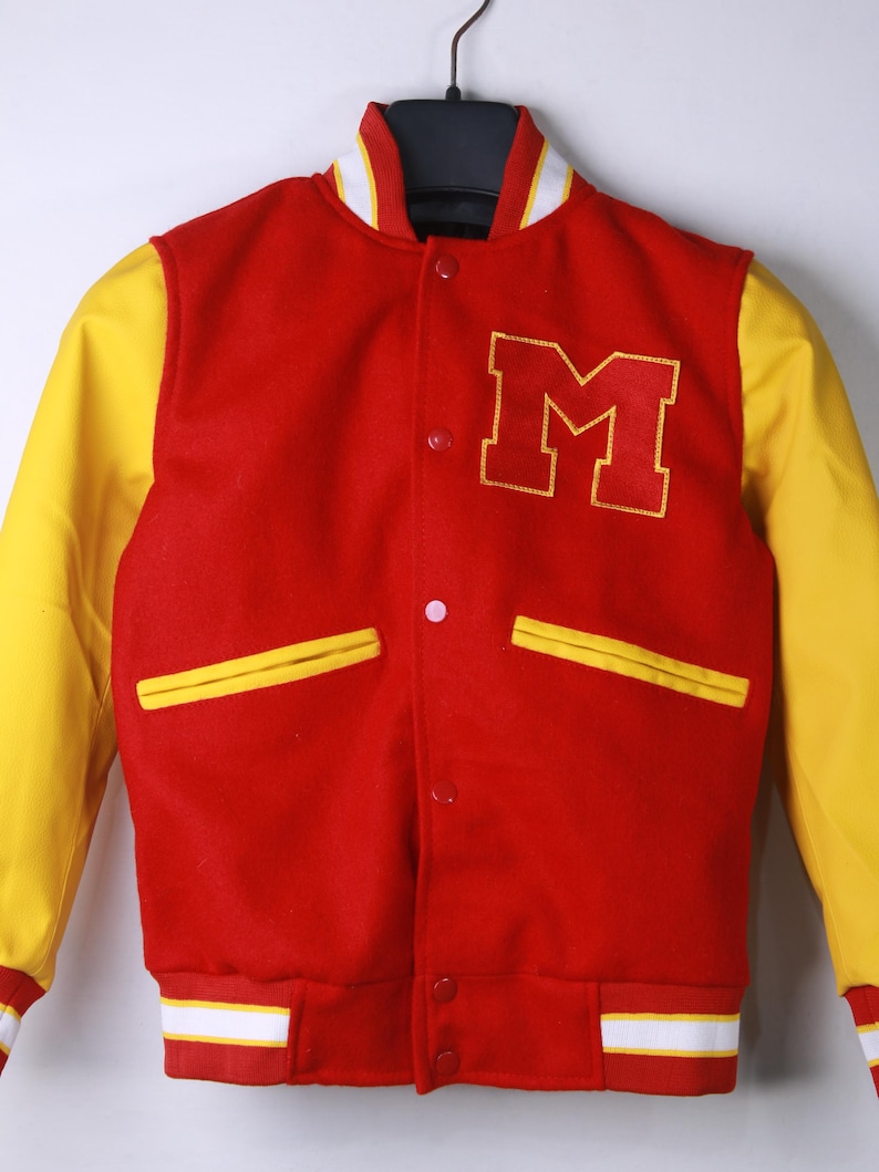 May include: A red and yellow varsity jacket with the letter "M" embroidered on the chest. The jacket features yellow sleeves, a white and yellow striped collar, and white and yellow striped cuffs. The jacket has two front pockets and a button-up closure.