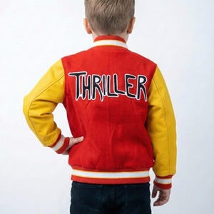 May include: Red and yellow varsity jacket with the word "THRILLER" in black and white lettering on the back. The jacket has yellow sleeves, a red body, and white and black striped trim. The jacket is worn with dark blue jeans.