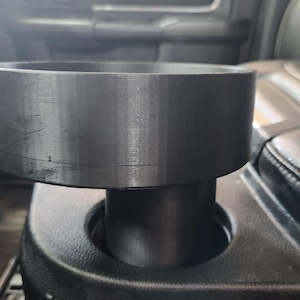 May include: A dark grey metal cup holder adapter, cylindrical in shape. Designed to fit into a vehicle's existing cup holder, it has a wide, flat top and a narrower base. The surface is smooth with a slight texture.