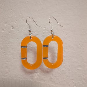Könnte beinhalten: Ein Paar orangefarbene, ovale Ohrringe mit silbernen Akzenten. Die Ohrringe haben einen Hakenverschluss und bestehen aus einem durchscheinenden Material. Ein auffälliges Schmuckstück.