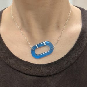 Könnte beinhalten: Halskette mit silberner Kette und einem durchscheinenden blauen ovalen Anhänger mit zwei weißen Balken. Der Anhänger hat eine leuchtende blaue Farbe, und die Halskette wird um den Hals getragen.