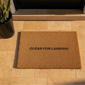 May include: A rectangular doormat made of brown coir fibre. The mat has the phrase "CLEAR FOR LANDING" printed in black. A black potted plant is visible in the upper left corner.