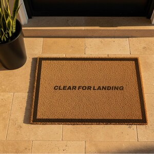 May include: A rectangular doormat made of natural coir fibre, featuring a brown background with a black border. The text "CLEAR FOR LANDING" is printed in black across the centre. A black planter with a green plant is visible in the background.