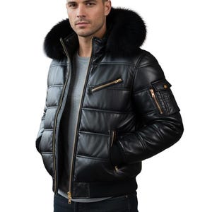 Peut inclure: Veste de bombardier en cuir noir avec capuche bordée de fourrure. La veste présente un design matelassé, plusieurs poches zippées et un ourlet côtelé. Les fermetures éclair et les accents dorés ajoutent une touche de contraste. La veste est portée sur un t-shirt gris.