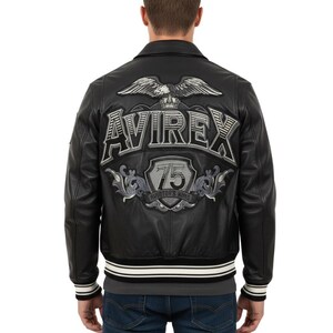 Könnte beinhalten: Schwarze Lederjacke mit weißem und schwarz gestreiftem Saum. Auf der Rückseite befindet sich ein großes gesticktes Design mit dem Wort "AVIREX", einem Adler und der Zahl "75".