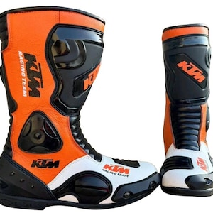Könnte beinhalten: Paar orange, schwarze und weiße KTM Racing Team Motorradstiefel. Die Stiefel haben ein hohes Design mit dem KTM-Logo und dem Text Racing Team. Die Stiefel sind aus Leder gefertigt und verfügen über Schutzeigenschaften.