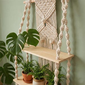 May include: Macrame plant shelf with two wooden shelves and hanging knotted rope supports. The shelf is adorned with green plants in terracotta pots and a clear glass vase. The macrame backdrop adds a bohemian touch to the decor.