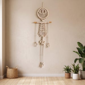 May include: Cream-colored macrame wall hanging with a geometric design. The wall art features a circular top, a wooden dowel, and hanging strands with knotted details. The piece is displayed on a neutral-toned wall, with a basket and plants in the background.