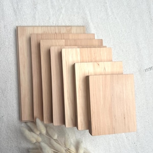 Puede incluir: Una colección de bloques de madera rectangulares de varios tamaños, dispuestos en un patrón escalonado. Los bloques tienen un tono de madera claro y natural y están apilados sobre una tela de color neutro. Un ramo de flores blancas secas y esponjosas está en primer plano.