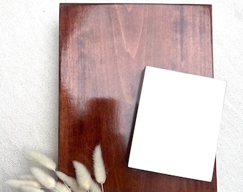 May include: A square, stained wooden block with a glossy finish, paired with a smaller, white square. A bundle of dried, fluffy, cream-colored flowers rests near the base. The wood has a rich, reddish-brown hue.