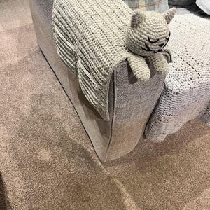 May include: A light grey crocheted cat armrest cover. The cat has a smiling face with closed eyes and small ears. The cover is draped over the arm of a grey sofa. The cat's tail hangs over the back of the sofa.