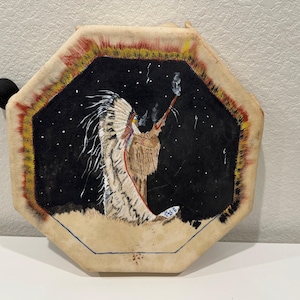 May include: An octagonal Native American drum featuring a painted scene of a figure in a feathered headdress against a starry night sky. The drum has a black handle and a beige, textured surface with red, yellow, and black painted edges.